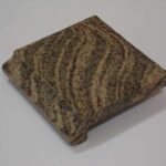 A small square sample block of tan and reddish-brown wavy-patterned granite featuring an ogee-style finished edge profile.