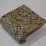 A small square sample block of tan, white, and black speckled granite featuring a rounded bullnose edge profile.
