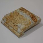 A small square stone material sample made of polished beige and white patterned marble or granite with a finished edge profile.