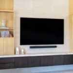Built-in entertainment unit with floating dark wood cabinets, light stone countertop, wall-mounted TV, soundbar, and light wood shelving units.