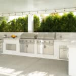 Covered outdoor patio kitchen with built-in stainless steel grills, white stucco bases, marble countertops, and string lights.