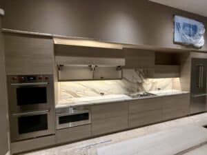 Modern luxury kitchen featuring light grey wood-grain cabinets, open upper lift-up doors, a dramatically veined marble backsplash, under-cabinet lighting, and stainless steel Wolf appliances.