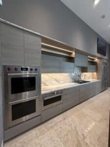 Modern luxury kitchen featuring grey wood-grain cabinets, a white and gold veined marble backsplash, under-cabinet LED strip lighting, and stainless steel Wolf appliances.