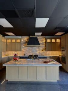 Symmetrical view of a luxury kitchen showroom display featuring light oak cabinetry, a large island with glowing backlit stone countertop, and a matching illuminated stone range wall