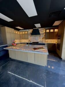 Kitchen showroom display featuring a large island with backlit stone countertop, light wood cabinetry, brass faucet, and a glowing stone backsplash behind a dark range hood.