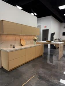 Modern kitchen showroom display featuring floating light oak cabinets, an illuminated white marble backsplash, and an attached waterfall island over a glossy epoxy floor.