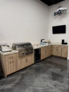 Outdoor kitchen showroom display featuring light wood L-shaped cabinetry, built-in stainless steel grill, mini fridge, white quartz countertop, and glossy epoxy floor.