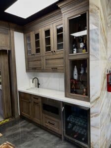 Angled view of a custom wood wet bar with glass display cabinets, white quartz countertop, black faucet, and an under-counter wine cooler.