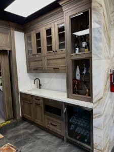 Angled view of a custom wood wet bar with glass display cabinets, white quartz countertop, black faucet, and an under-counter wine cooler.