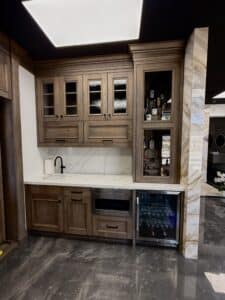 Custom medium-wood wet bar featuring glass-front display cabinets, white quartz countertops with delicate grey veining, a matte black faucet, and a stainless steel beverage fridge.