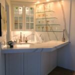 Elegant corner wet bar featuring thick white stone countertops with bold grey veining, floating glass shelves, and illuminated glass-front cabinetry.
