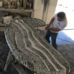 Worker hand-finishing the curved edge of a dark grey and white veined stone island top in a workshop.