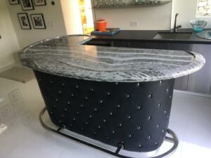 A custom curved home bar with a polished gray-and-white marbled granite top, black tufted leather base, and chrome footrail.