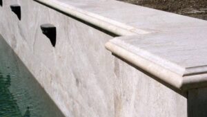 Travertine stone pool coping with bullnose edges lining a retaining wall with decorative dark water spouts.