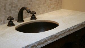 A dark bronze faucet and undermount sink set into a beige stone vanity countertop with a chiseled, chiseled edge.