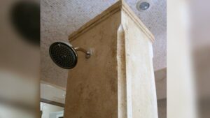 A round metal shower head mounted directly onto a beige travertine stone column in a tiled shower enclosure.