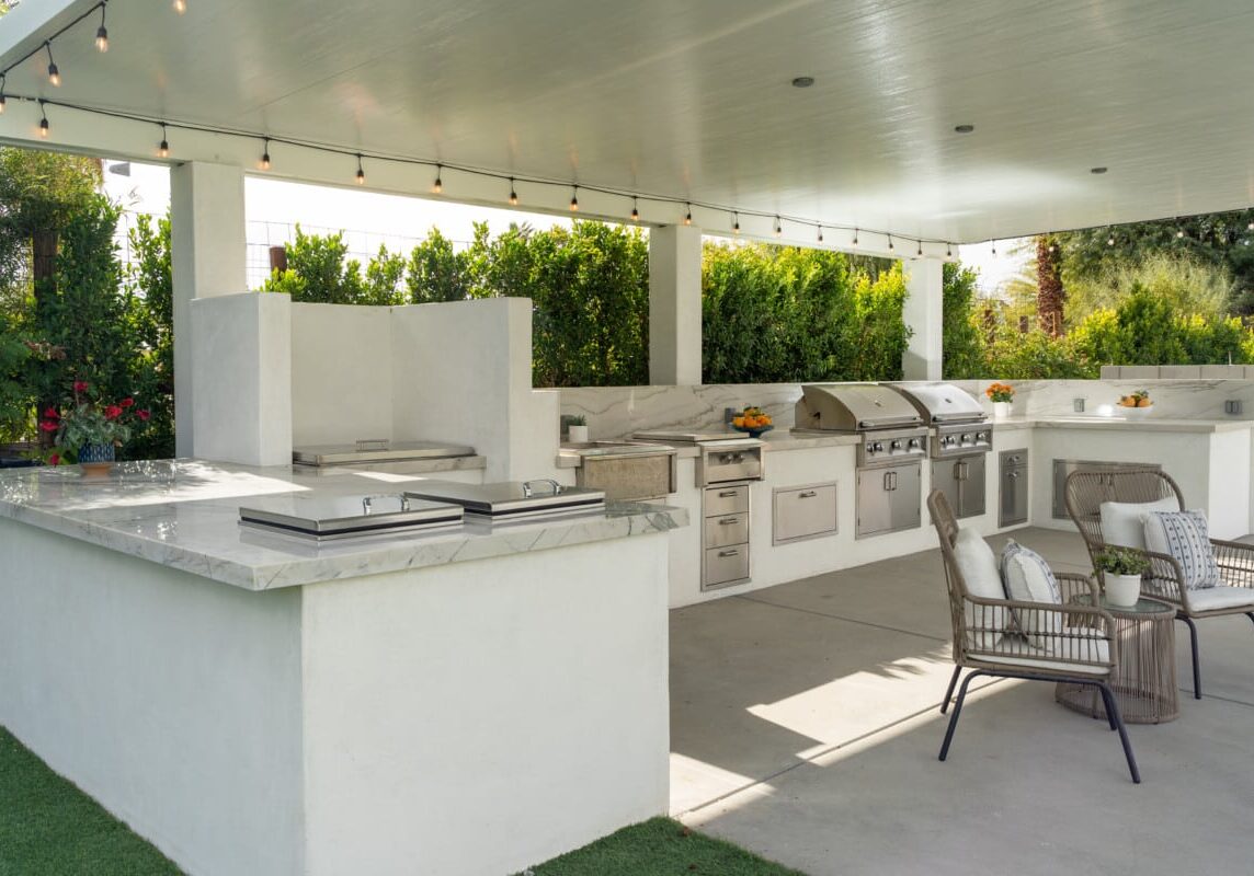 Covered outdoor patio featuring a spacious kitchen with white stucco cabinetry, marble countertops, built-in stainless steel grills, and woven lounge seating.