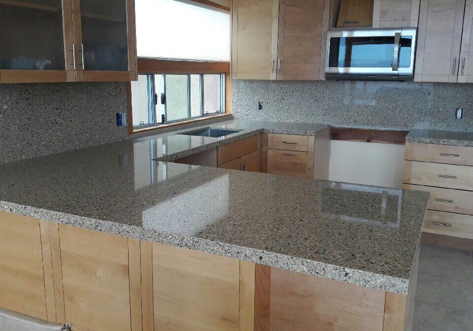 Polished gray and black speckled granite countertops and matching full-height backsplash in a kitchen with light wood cabinetry.