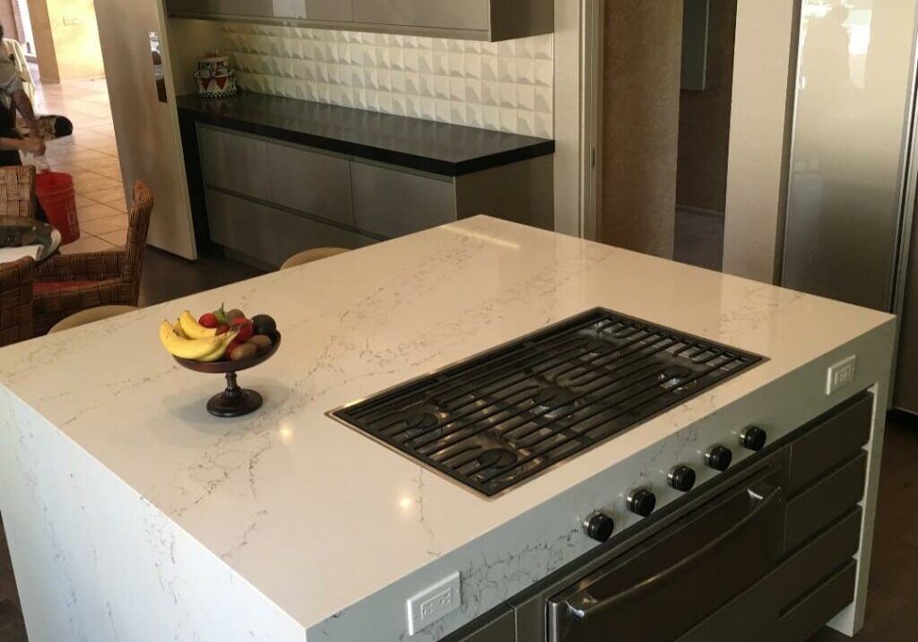 Modern kitchen featuring a large white marbled quartz island with a waterfall edge, built-in cooktop, and a bowl of fruit.